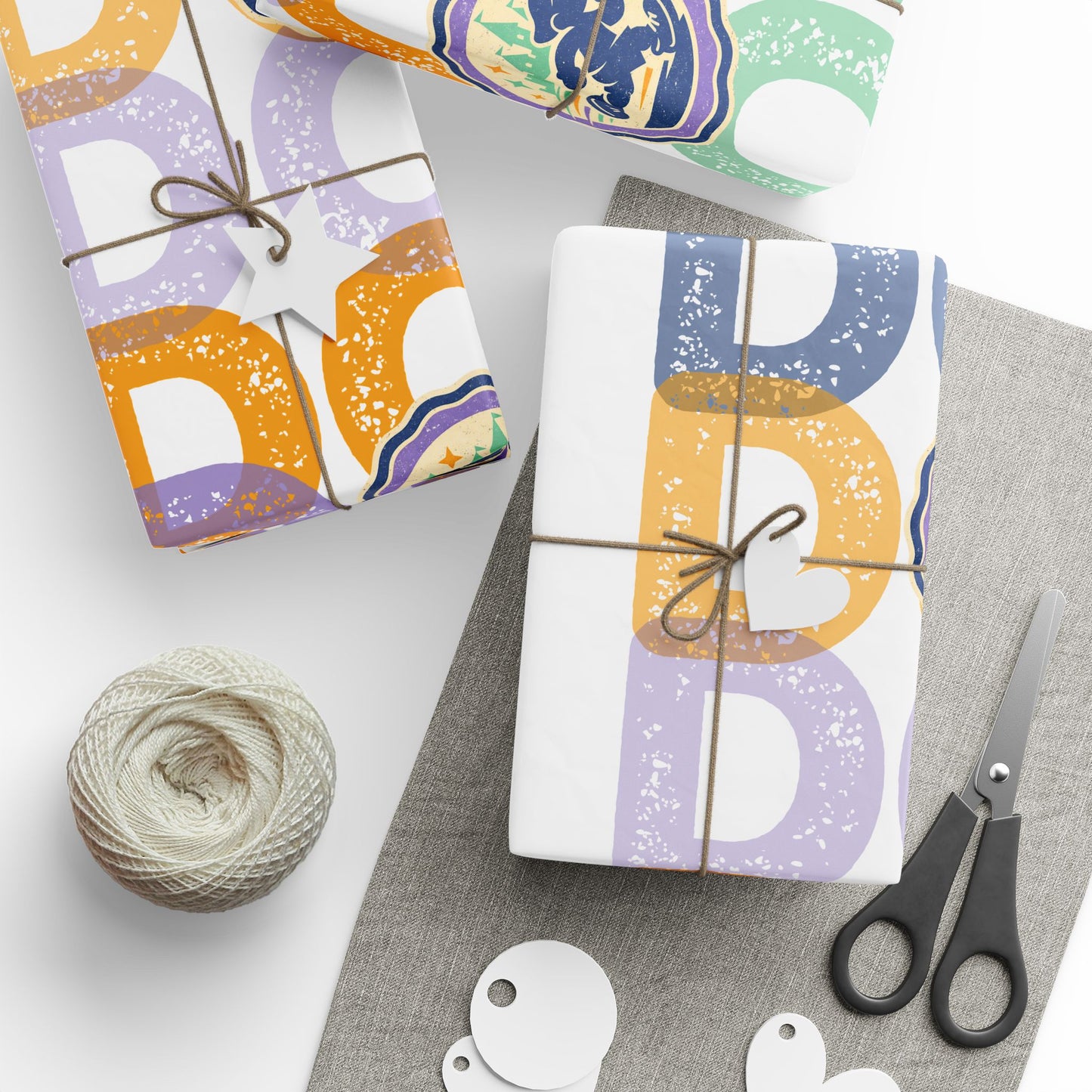 Three gift boxes wrapped in white wrapping paper with large distressed pastel letters (orange, purple, and blue) are arranged on a light gray surface with twine, scissors, string, and gift tags.