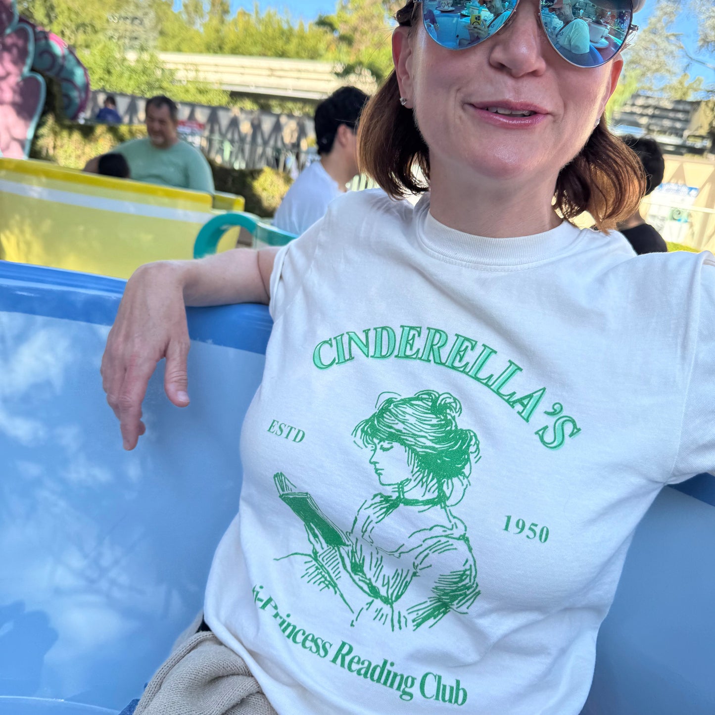woman with sunglasses wearing a cinderella's anti-princess reading club tee. ivory tee with green graphic and lettering on a tea cup ride in disneyland