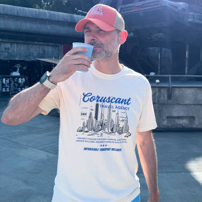 Man wearing a 'Coruscant' t-shirt drinking from a cup outdoors with a structure and trees in the background.