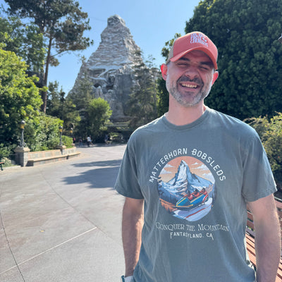 Man wearing a cap and matterhorn disneyland t-shirt with a mountain design, standing in front the matterhorn ride