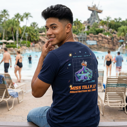 Man wearing a navy blue t-shirt with a graphic design and text, sitting by a pool area with people and chairs in the background.