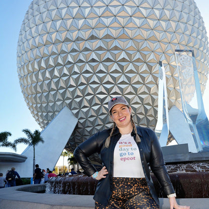 Person posing in front of a large, geometrically shaped structure with palm trees in the background.