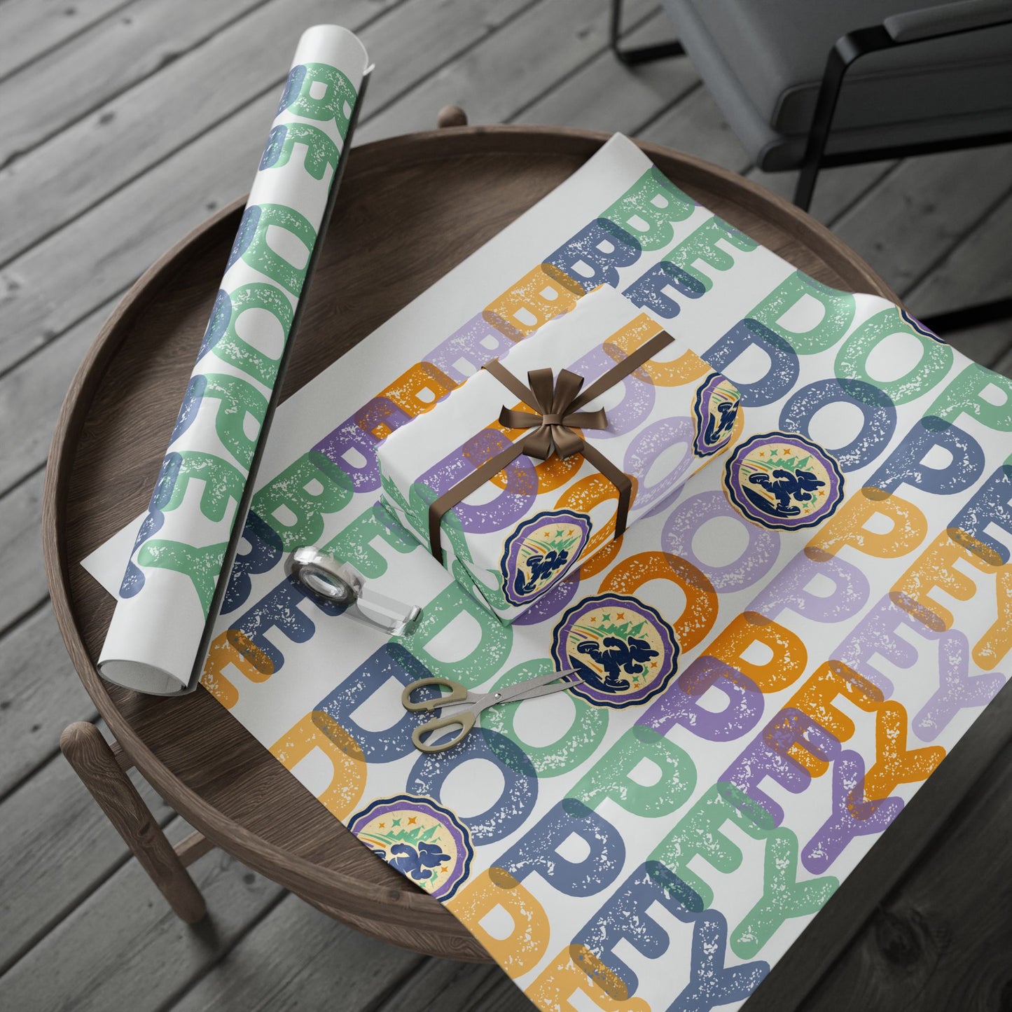 A roll and flat sheet of wrapping paper with a colorful 'BE DOPEY' typographic pattern in green, orange, purple, and blue, styled on a round wooden tray with scissors and a small wrapped gift on a gray deck.