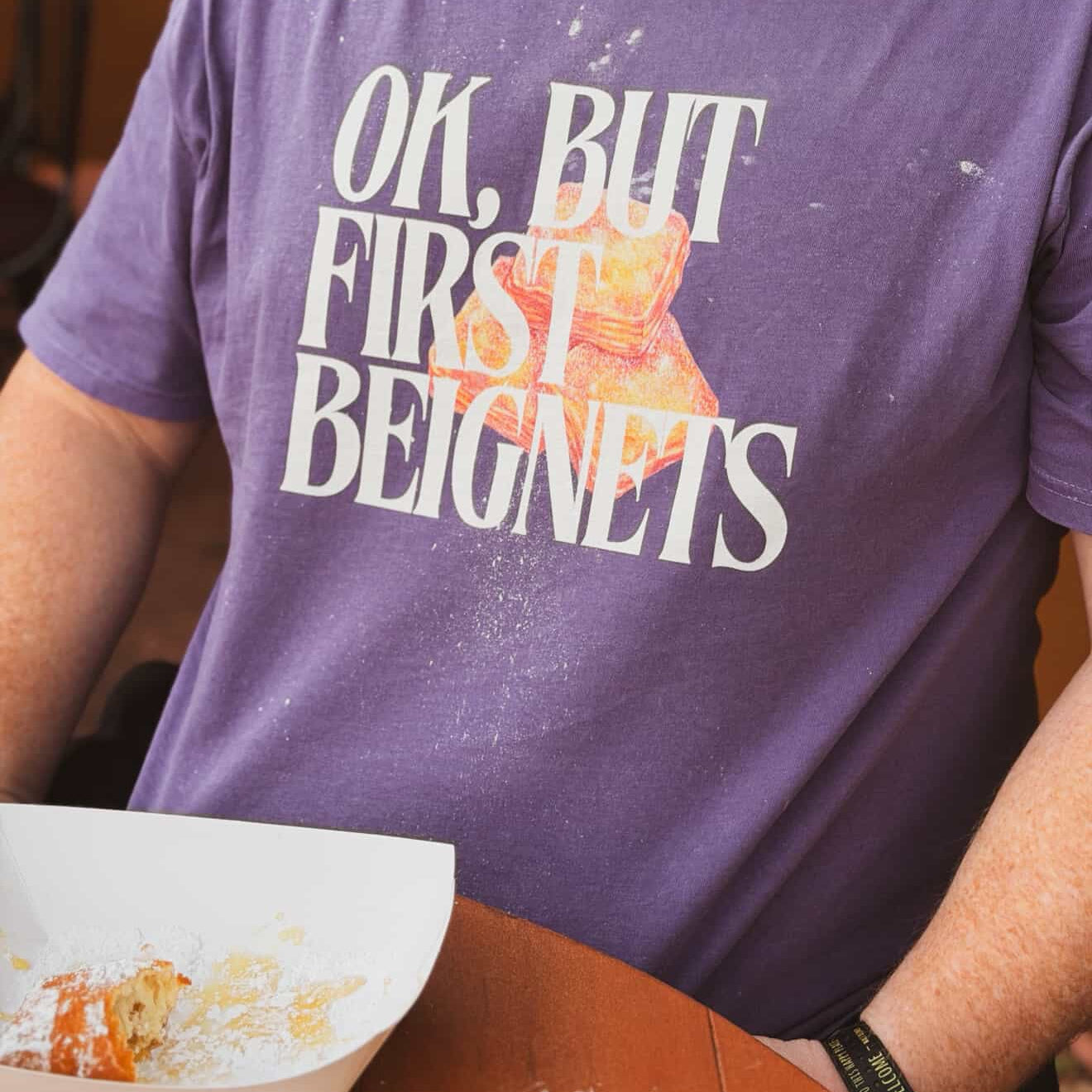 Person wearing a purple t-shirt with a graphic and text, sitting at a table with a plate of food.