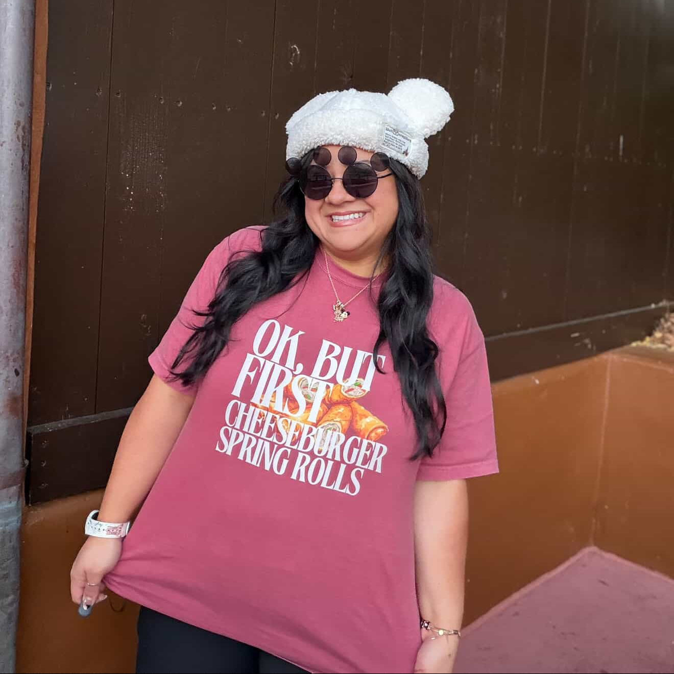 Person wearing a pink t-shirt with text, sunglasses, and a white beanie standing against a brown wall.