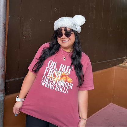 Person wearing a pink t-shirt with text, sunglasses, and a white beanie standing against a brown wall.