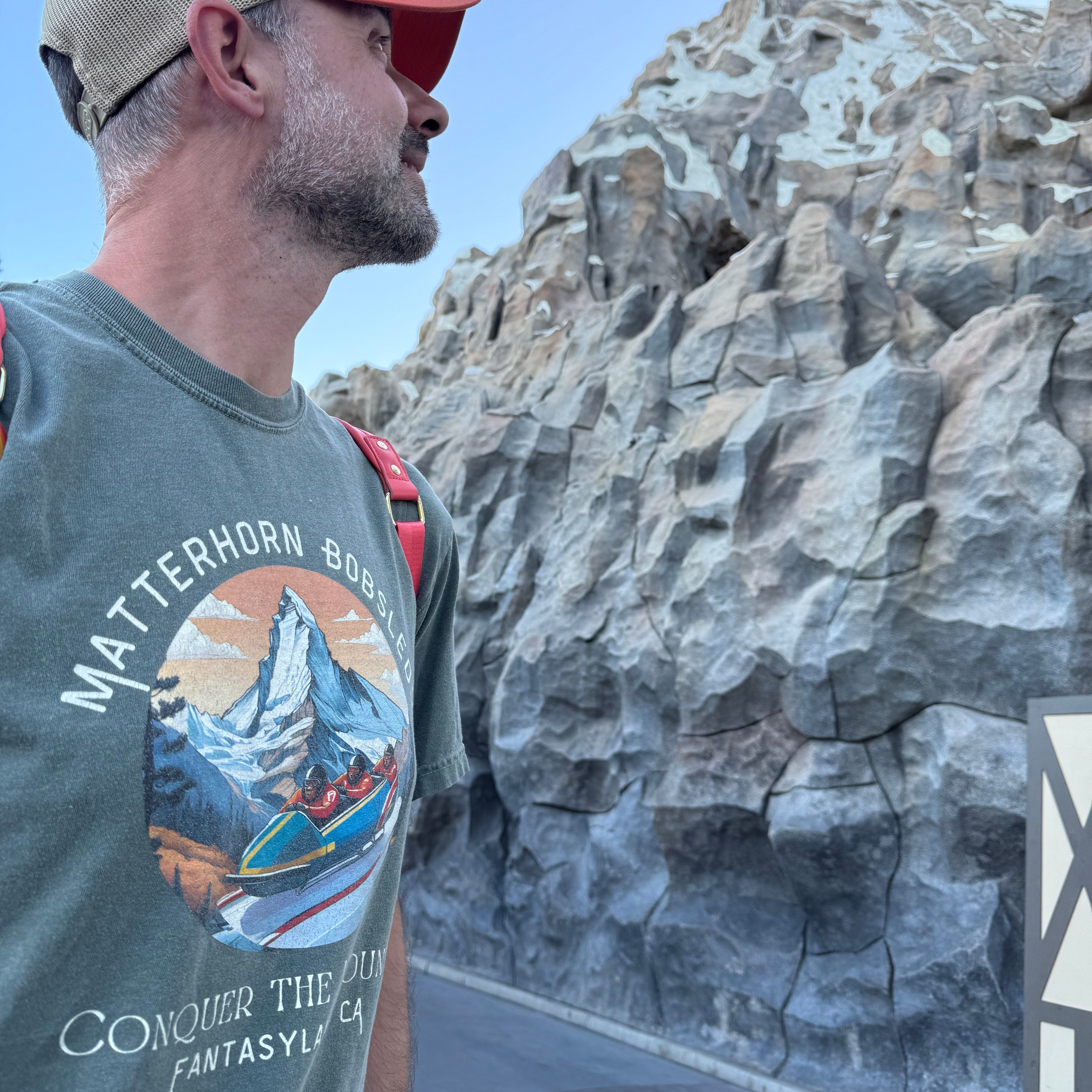 Man wearing a cap and matterhorn disneyland t-shirt with a mountain design, standing in front the matterhorn ride