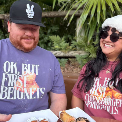 Two people sitting outdoors with food, wearing matching t-shirts with humorous text.