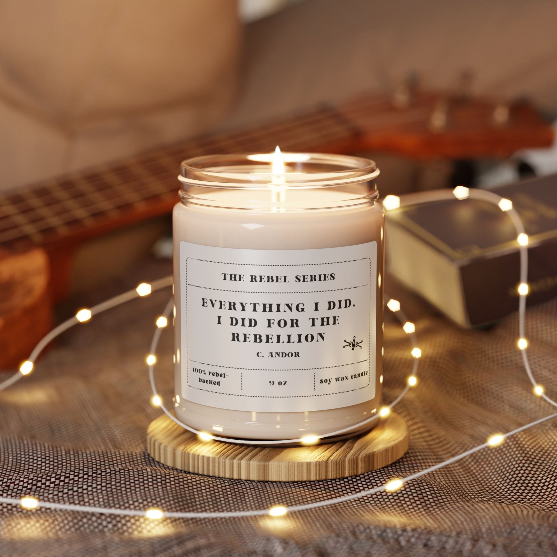 Lit candle in a jar with a label on a wooden coaster, surrounded by string lights and a guitar in the background.